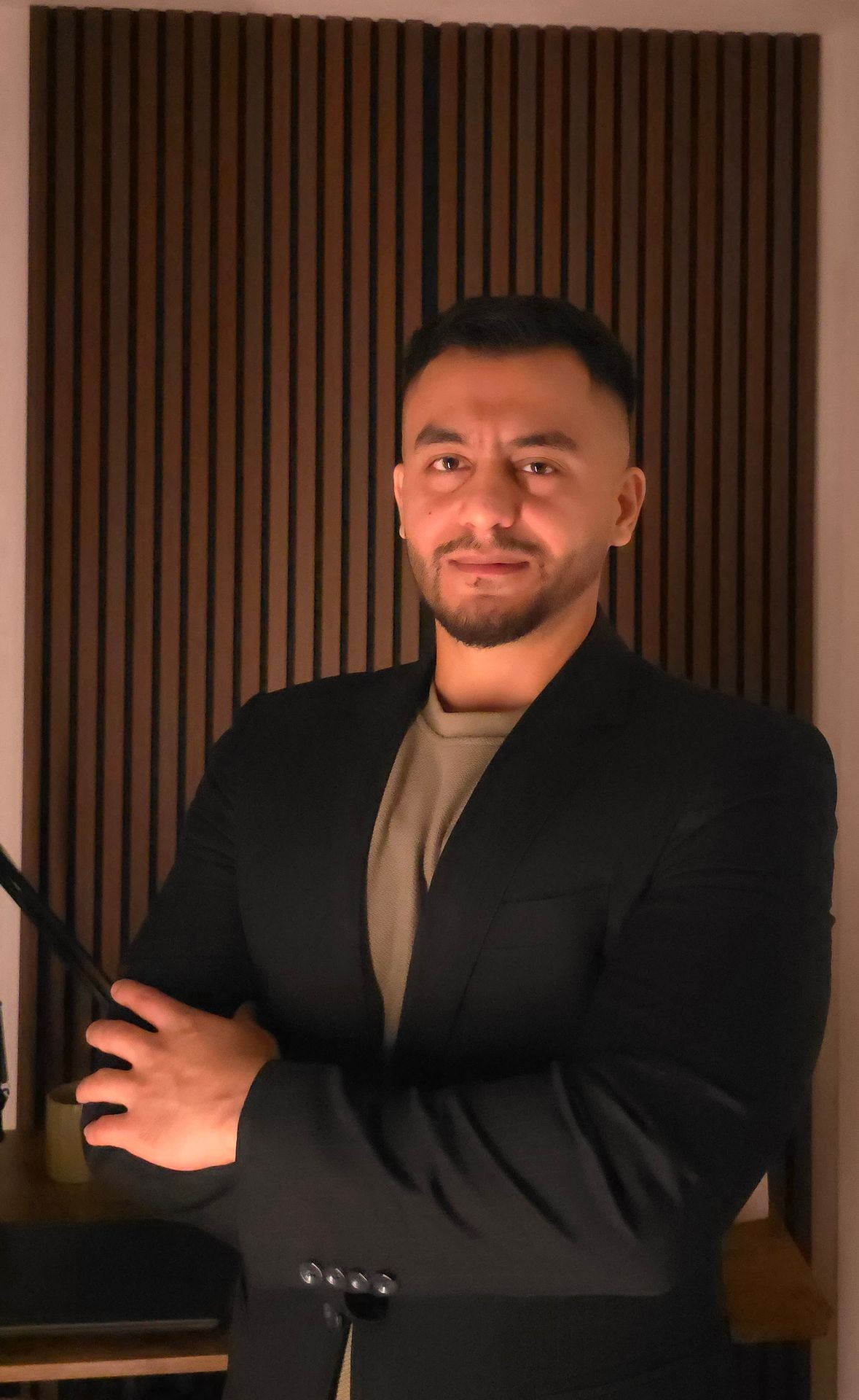 Person wearing a black blazer and light-colored shirt standing in front of a wooden slat wall.